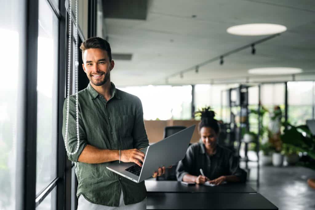 Jeune homme souriant tenant un ordinateur portable dans un bureau moderne et lumineux.
