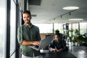 Jeune homme souriant tenant un ordinateur portable dans un bureau moderne et lumineux.