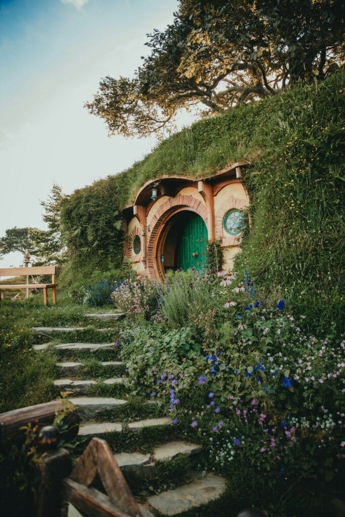 Trou de Hobbit iconique avec porte ronde verte et toit de gazon, entouré de marches en pierre et d'un jardin anglais luxuriant.
